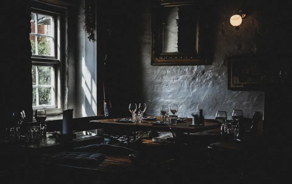La salle du restaurant dans la pénombre, tables dressées près de la fenêtre